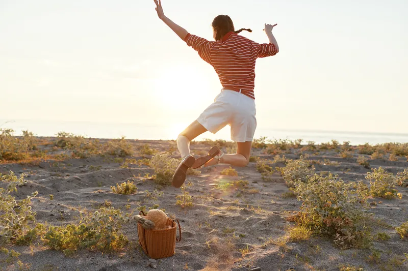 A happy young woman jumping for joy on a sandy beach during sunset. Lifestyle concept of freedom, leisure, and summer vacation outdoors with a picnic basket in the warm light.