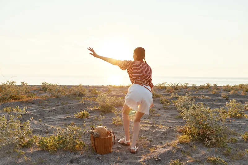 Young woman reaches towards the sunset on a sandy beach during summer vacation. She wears casual clothing near a picnic basket, enjoying the warm light in a beautiful coastal lifestyle scene.